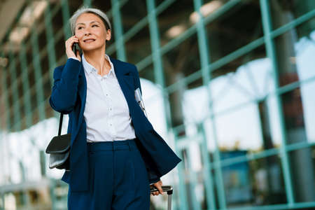 Grey asian woman talking on cellphone while walking with suitcase at airport parkingの写真素材