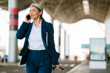 Grey asian woman talking on cellphone while walking with suitcase at airport parkingの写真素材