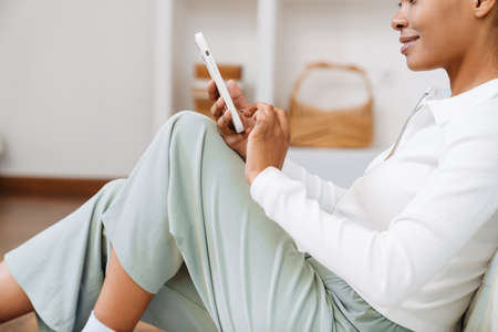 Young black woman using cellphone while sitting on floor at home