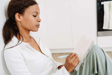 Young black woman writing down notes while sitting on floor in kitchen at home