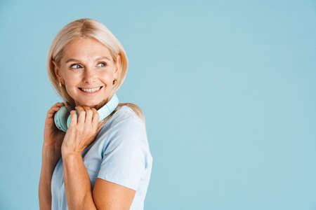 Mature blonde woman with headphones smiling and looking aside isolated over blue background