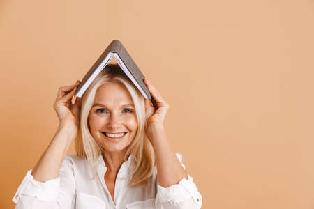 Mature woman covering her head and smiling at camera isolated over beige background