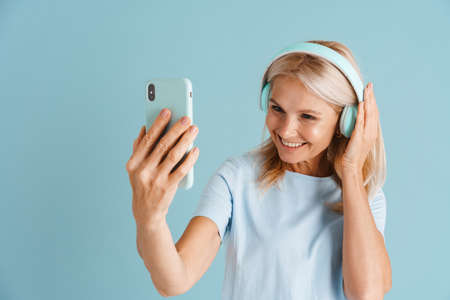 Mature blonde woman in headphones taking selfie on cellphone isolated over blue background