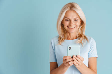 Mature blonde woman wearing t-shirt smiling and using cellphone isolated over blue background