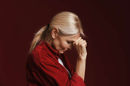 Mature blonde woman wearing shirt propping up her head isolated over red backgroundの写真素材