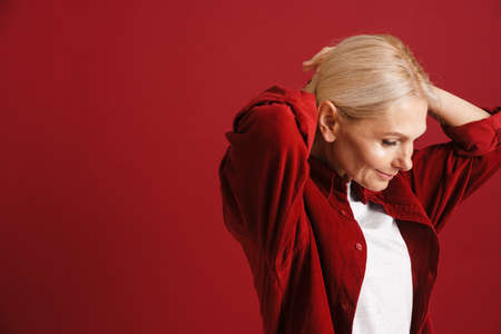 Mature blonde woman wearing shirt smiling and doing hairstyle isolated over red background