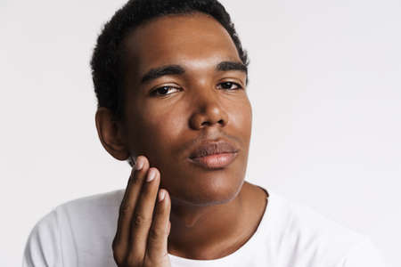 Young black man looking at camera while touching his skin isolated over white wallの写真素材
