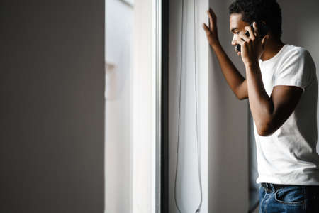 Young black man talking on cellphone while looking at window indoors