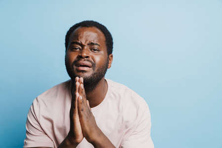 Black bristle man crying while holding his palms together isolated over blue background
