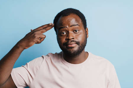 Black confused man in t-shirt making shot gesture isolated over blue background