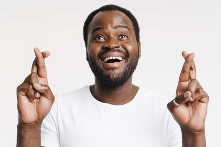 Black bristle man smiling while holding fingers crossed for good luck isolated over white background