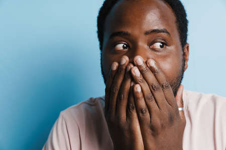 Black bristle man expressing surprise and covering his mouth isolated over blue backgroundの写真素材