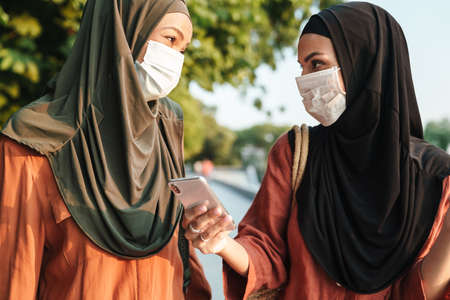 Multiracial muslim women in face masks using cellphone while walking together outdoorsの写真素材