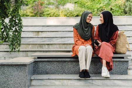Multiracial muslim women talking and smiling while sitting on bench outdoorsの写真素材
