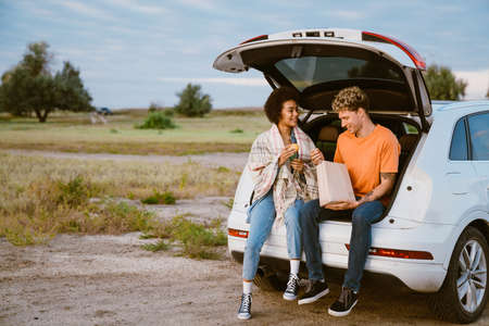 Young multiracial couple eating peach while sitting in trunk during car trip