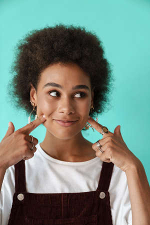 Young black woman smiling and pointing fingers at her cheeks isolated over blue backgroundの写真素材