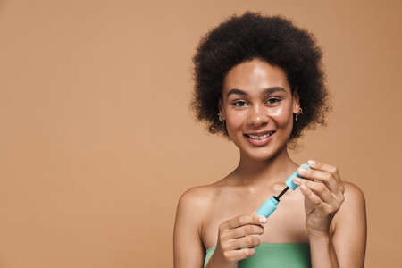 Black brunette woman smiling while posing with mascara isolated over beige background