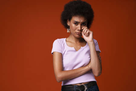 Young black woman wearing earrings frowning while wiping her tears isolated over brown background