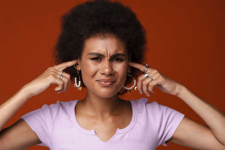 Young black woman wearing earrings frowning while plugging her ears isolated over brown background