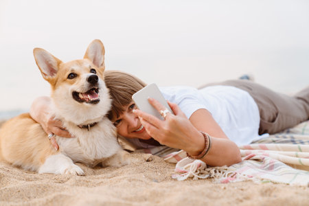 Senior woman taking photo on cellphone while resting with her dog on sandy beachの写真素材