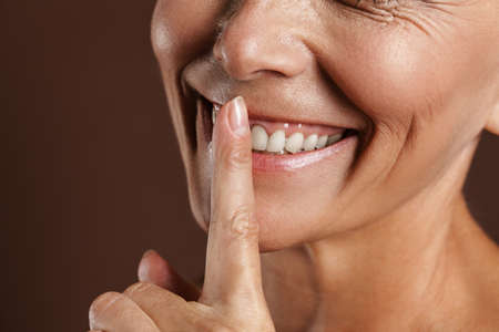 Senior shirtless woman smiling while showing silence gesture isolated over brown wall