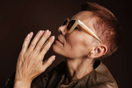 Senior ginger woman in sunglasses gesturing and looking aside isolated over brown wall