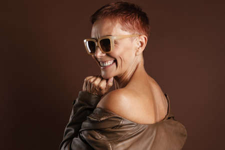 Senior ginger woman smiling while posing in leather jacket isolated over brown wall