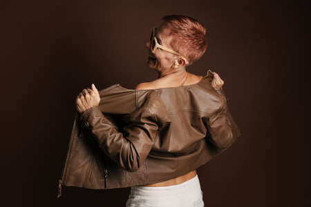 Senior ginger woman smiling while posing in leather jacket isolated over brown wallの写真素材