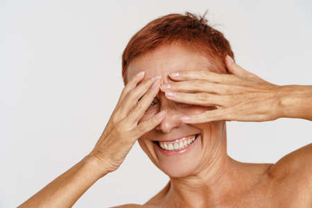 Senior shirtless woman with short hair smiling while covering her eyes isolated over white backgroundの写真素材