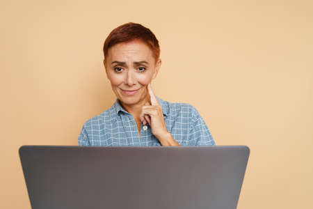 Senior ginger woman smiling while posing with laptop isolated over beige wall