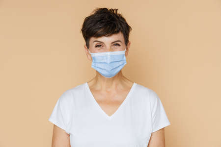 Mature european woman wearing t-shirt posing in face mask isolated over beige backgroundの写真素材
