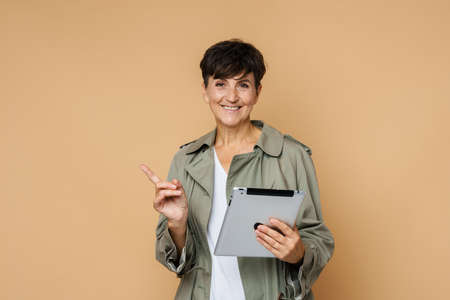 Mature european woman pointing finger aside while using tablet computer isolated over beige...