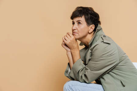 Mature european woman looking aside while sitting on chair isolated over beige backgroundの写真素材