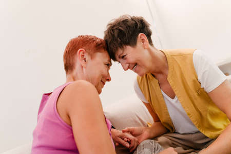 Mature lesbian couple hugging and smiling at each other at homeの写真素材