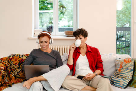 Mature lesbian couple using laptop and drinking coffee on sofa at homeの写真素材