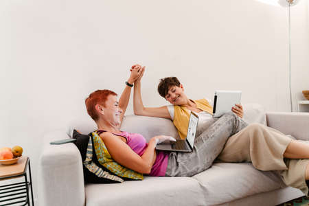 Mature lesbian couple holding hands while using gadgets on sofa at homeの写真素材