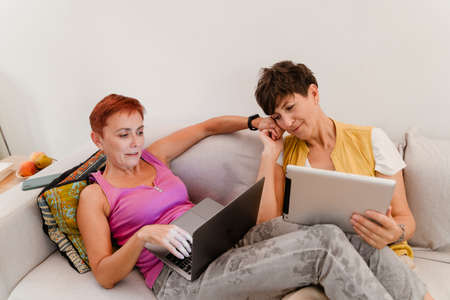 Mature lesbian couple holding hands while using gadgets on sofa at homeの写真素材