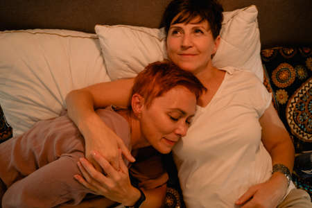 Mature lesbian couple hugging and smiling while lying in bed at homeの写真素材
