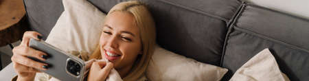 Young blonde woman using mobile phone while lying in bed after sleep at homeの写真素材