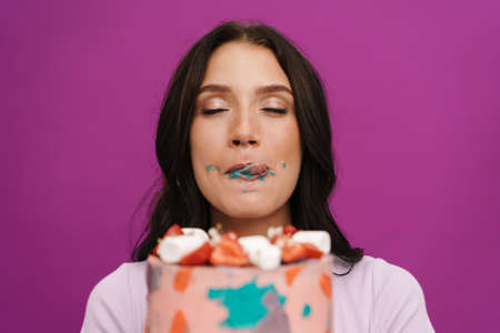 Young brunette woman wearing t-shirt eating birthday cake isolated over purple wallの写真素材