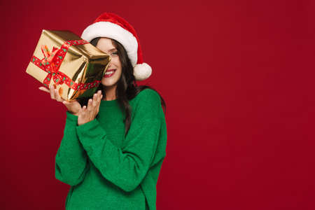 Young woman in Christmas hat smiling while posing with present box isolated over red wall