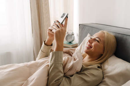Young blonde woman using mobile phone while lying in bed after sleep at home