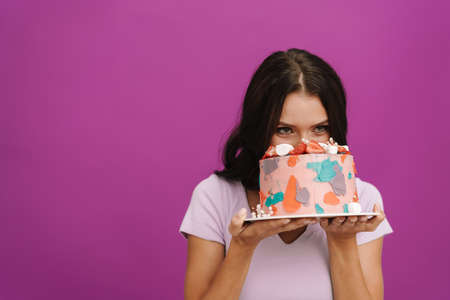 Young brunette woman wearing t-shirt eating birthday cake isolated over purple wallの写真素材