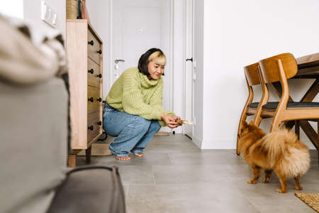 Asian girl smiling and feeding her dog while squatting at homeの写真素材