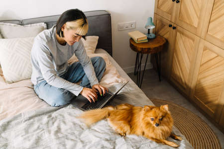 Asian girl using laptop while sitting with her dog on bed at homeの写真素材