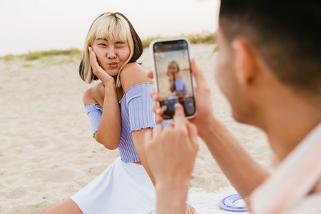 Young man taking photo of his girlfriend during picnic on summer beachの写真素材