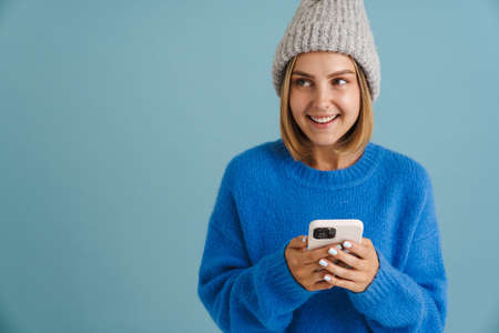 Young blonde woman wearing knit hat smiling and using cellphone isolated over blue background