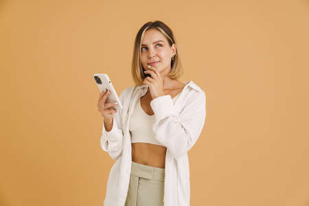 Young blonde woman looking upward while using mobile phone isolated over beige background