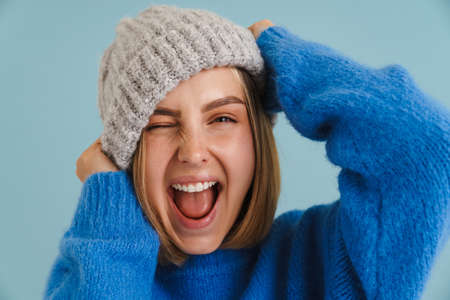 Young blonde woman wearing knit hat smiling and winking at camera isolated over blue backgroundの写真素材