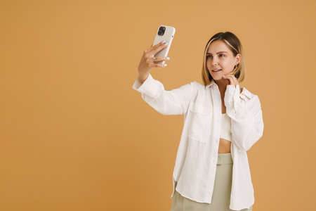 Young blonde woman taking selfie photo on mobile phone isolated over beige background